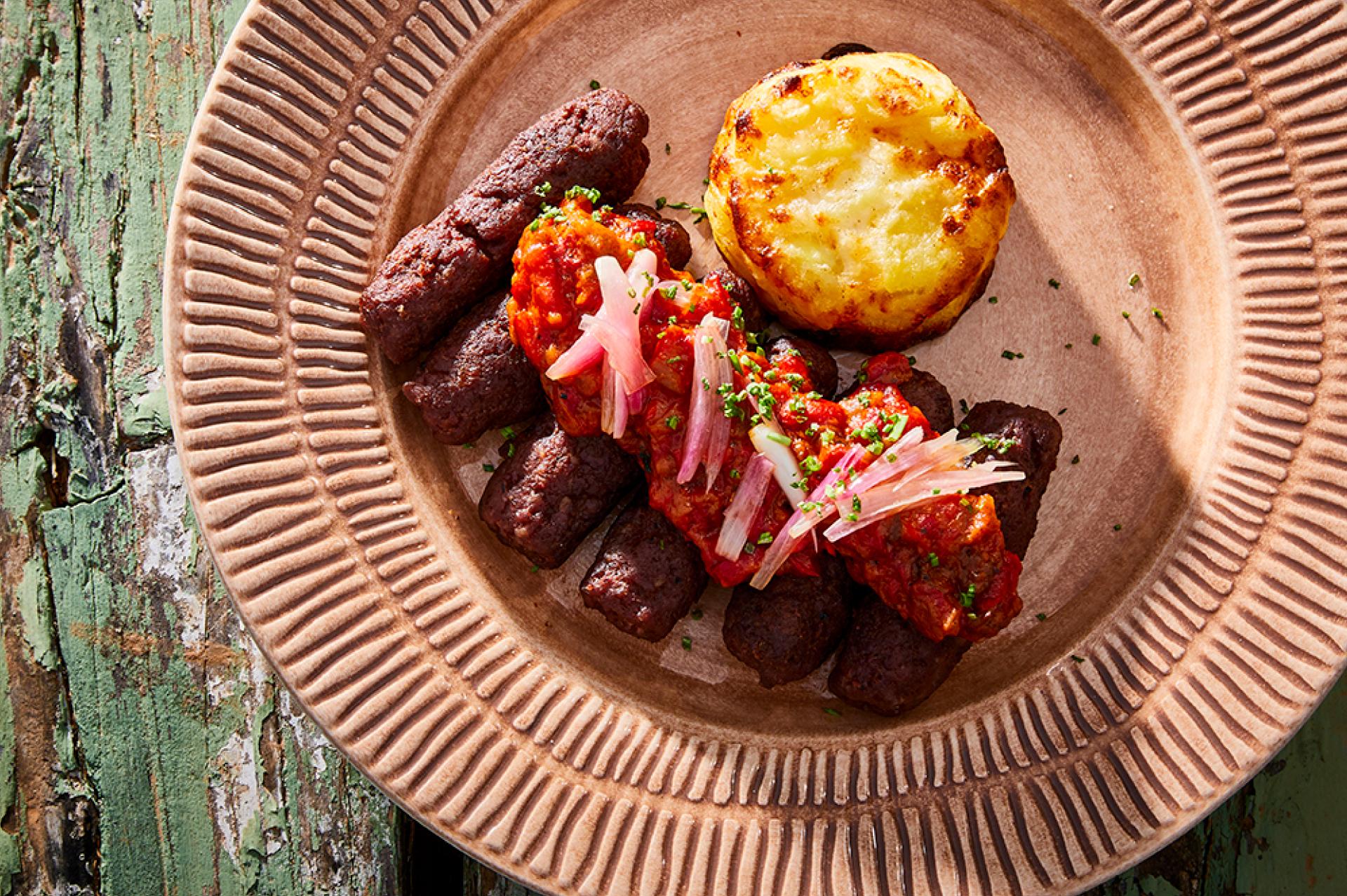 cevapcici auf grünem holztisch und rosa teller serviert mit aviko gratin