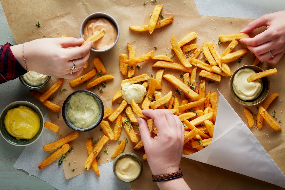 Pommes auf braunem Papier ausgebreitet, mehrere Leute essen Pommes mit verschiedenen Mayonaise Variationen