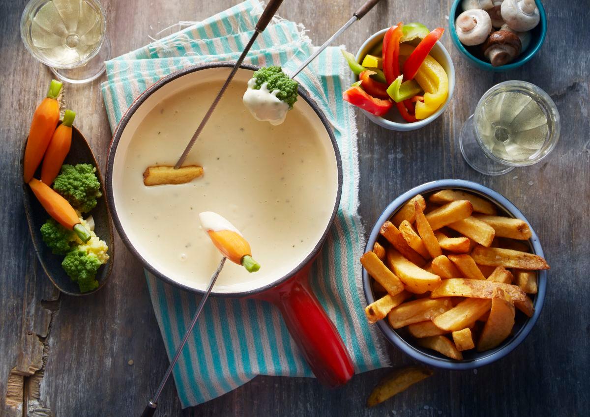 Käse fondue in welches Pommes und eine Karotte gedippt wird auf Tisch mit beilagen serviert