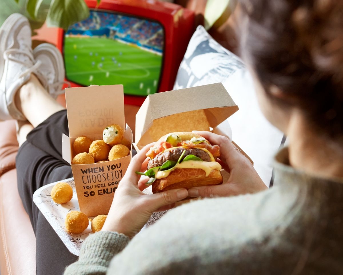 Mann isst bestellten burger mit pommes aus der verpackung vor dem fernseher