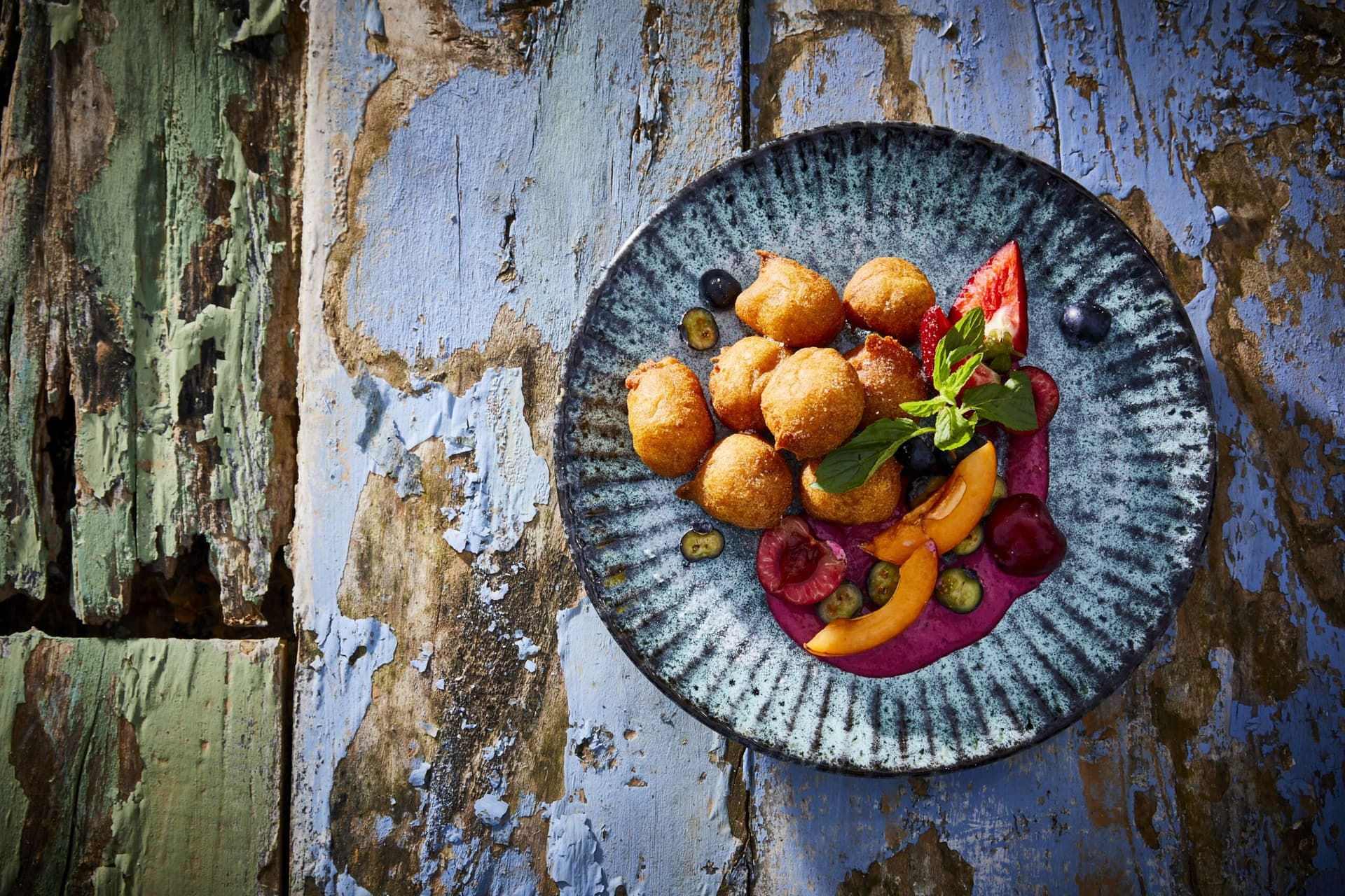 Fritule serviert auf blauem tisch und blauem teller neben früchten und pinker sosse